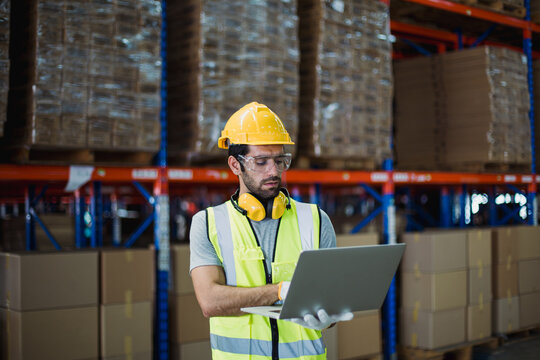 Warehouse logistics coordinator wearing safety gear using laptop to manage transportation schedules and cargo operations in a busy industrial distribution center.