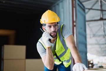 Logistics worker wearing safety gear using walkie talkie inside warehouse to coordinate freight...
