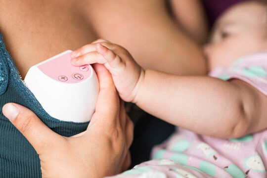 Mother using a wearable electric breast pump while breastfeeding her baby. Real moment of modern lactation, milk expression, and maternal care at home