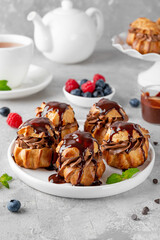 Profiteroles with chocolate cream, fresh berries and chocolate glazed on a plate on a gray concrete background. A delicious French dessert.
