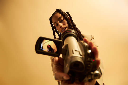Young adult Caucasian woman filming with handheld video camera, looking down at device with focused expression, camera screen displaying her reflection, beige background
