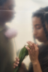 Young Black man standing beside young Caucasian woman holding pink flower, woman gently touching petals while man looking at her, both partially blurred by translucent surface