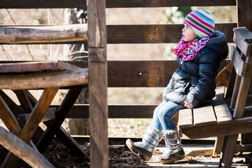 Little girl wearing winter clothes sitting on wooden bench in farmhouse garden