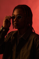 Young adult Black man adjusting sunglasses while facing camera, wearing casual jacket, standing...