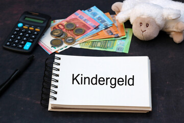 Das Wort Wort Kindergeld auf einen Notizblock geschrieben mit Taschenrechner, Geldscheinen und einem Teddybär