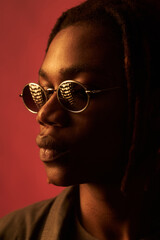 Young adult Black man wearing round sunglasses standing in profile against plain background, lips...