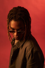 Black young adult man wearing sunglasses looking down with serious expression against plain red...