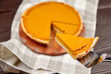 Delicious pumpkin pie on wooden board with slice being served on metal spatula, showcasing rich...