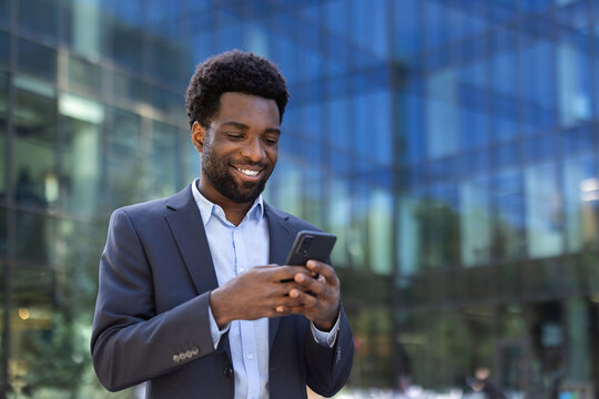 An African-American businessman smiles as he looks at his phone while outside a modern office building.