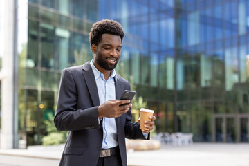A smiling businessman in a suit checks his phone while holding a coffee cup outside a modern office...