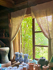 Window in country house with curtains made of coarse burlap, clay products on windowsill. Potters workshop. View from the window.