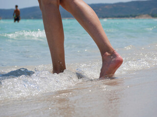 Close-up of female legs walking barefoot along the beach with sea waves. Wellness, travel and lifestyle concept