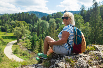 Active senior woman with a backpack and red cap sitting peacefully on a rocky cliff overlooking a...
