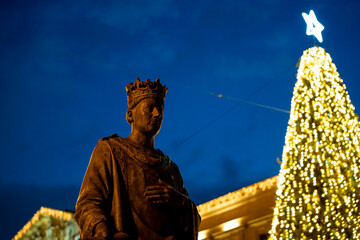 Statue of a regal figure with a brightly lit Christmas tree in Pamplona during the evening...