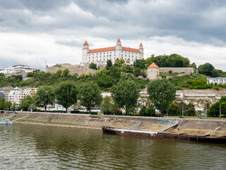 Naklejka premium Panoramic view of Bratislava Castle above the Danube River with trees and buildings in the foreground under dramatic clouds.