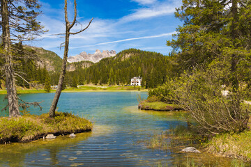 Picturesque Lago Misurina with its clear waters, framed by pine trees and distant rocky mountains, featuring a charming building on the far shore under a blue sky.