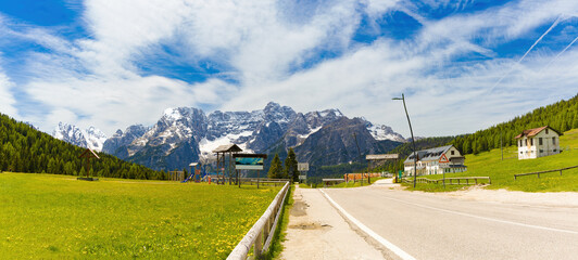 Panoramic view of the majestic snow-capped Cristallo mountain range in the Dolomites, towering over green alpine meadows, a road, and scattered buildings under a bright sky.