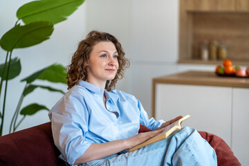 Mindful woman enjoys reading book at home, digital detox. Serene girl sits on sofa holding novel,...