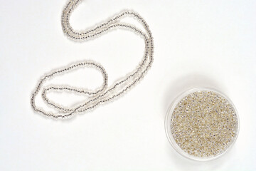 A plastic container full of tiny silver-lined seed beads is positioned on a light background,...
