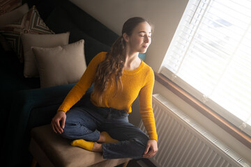 Young woman meditating in lotus pose on chair near window at home, eyes closed, wear casual...