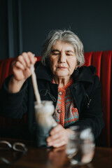 Elderly woman mixing a drink during a relaxing moment at a cafe, wearing a colorful knitted sweater...