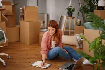 A woman sits on the floor of her new home surrounded by cardboard boxes. She focuses on writing in...