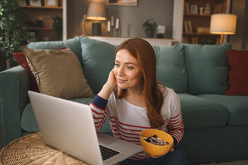 A woman sits comfortably on a plush couch, engaging with content on her laptop while holding a bowl...