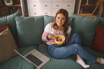 A woman sits relaxed on a soft couch, smiling as she eats a bowl of colorful cereal. A laptop is...