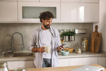 A man is sipping coffee and looking at his smartphone in a stylish kitchen filled with natural...