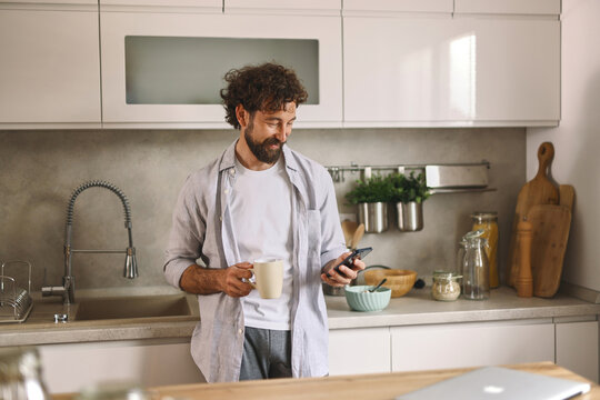 A man is sipping coffee and looking at his smartphone in a stylish kitchen filled with natural light. The space features modern appliances and simple decor, creating a cozy atmosphere.
