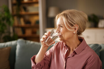 In a warm living room, a senior woman sips water from a glass, reflecting on her day while seated...