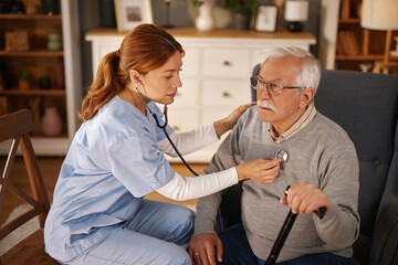 A female healthcare provider in blue scrubs uses a stethoscope to check the heart of an elderly male patient. The patient sits in an armchair, using a cane, in his home.
