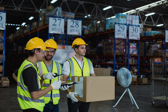 Warehouse staff in safety helmets and vests collaborating in logistics center, managing packages with checklist and scanner in modern storage facility.  Logistics teamwork concept,