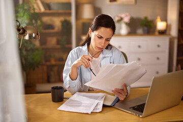 A woman appears focused as she sits at a wooden desk cluttered with papers, analyzing her finances....