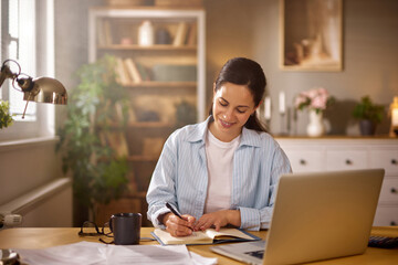A woman sits at her desk in a home office, writing in a notebook with a laptop in front of her. She enjoys a cup of coffee, surrounded by plants and cozy decor.