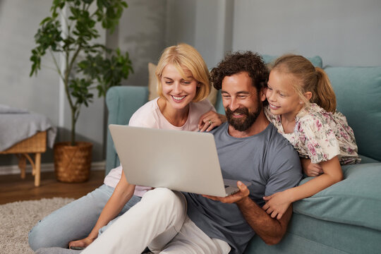 A family sits on a soft rug in their living room, sharing joyful moments as they gather around a laptop. The warmth of the afternoon sun creates a comfortable atmosphere.