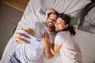 Smiling couple lies on the bed, playfully taking a selfie together. They appear relaxed and happy, enjoying a private afternoon in their cozy bedroom space.