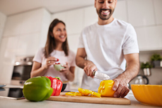 In a modern kitchen, a smiling couple engages in cooking, with one person slicing a yellow bell pepper and the other holding a bowl, radiating joy and togetherness.