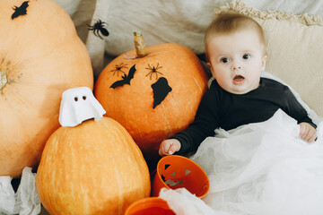 Happy Halloween. Cute happy baby in ghost costume sitting with jack o lantern bucket, pumpkins,...
