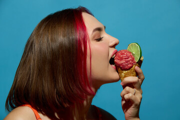Young woman with red hair biting lime and meat ice cream cone with curious expression. Concept of food paradox, surreal pop art, culinary experiments and provocative beauty ads.