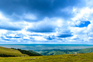 Rolling green hills descend into a wide valley with a distant lake shimmering under a dramatic sky...