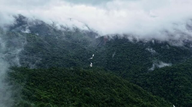 An epic drone view of a cascading waterfall lush mountains and forests topped with misty clouds