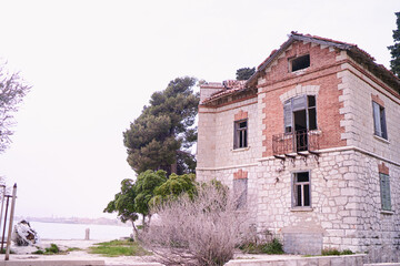 An abandoned, dilapidated stone house by the seaside, surrounded by trees and a cloudy sky.