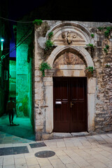 A historic stone building with an arched doorway, adorned with greenery, is illuminated at night. A...