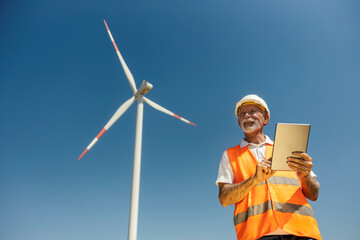 Low angle view of senior environmentalist with tablet standing near wind turbine.