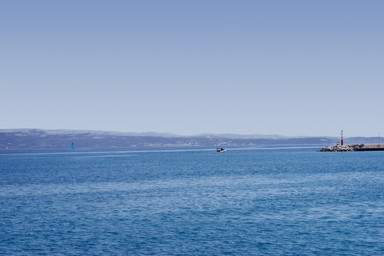 A serene seascape with a calm ocean, distant sailboats. The horizon is visible with a small lighthouse and marina on the right side of the image.