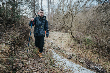 A man wearing outdoor gear walks along a nature trail surrounded by trees and a stream. The scene...