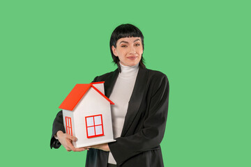 Woman in a black robe holding a toy house with red roof against a bright green background during a...