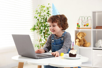 Birthday celebration. Little boy with cupcake having video call via laptop at table indoors