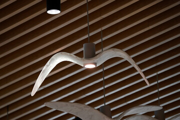 Unique ceiling lights shaped like flying birds hang from a wooden slatted ceiling. Each light emits a soft glow from the bird's body, creating an artistic and natural ambiance indoors.